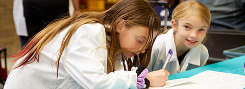 Picture of two girls wearing lab coats. One is writing something with a pen.